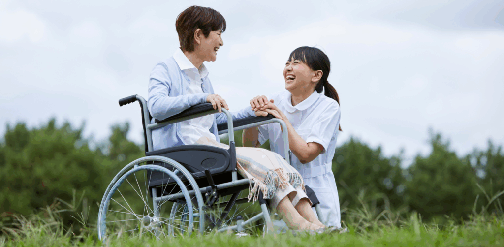 Smiling nurse kneels beside a woman in a wheelchair outdoors, sharing a joyful moment