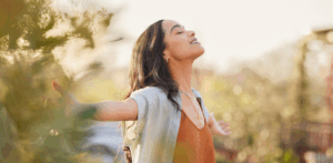 Young woman outdoors with arms outstretched, eyes closed, enjoying peaceful sunlight