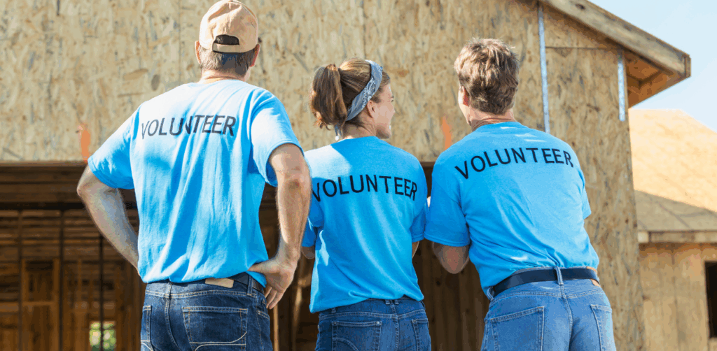 Three volunteers in matching blue shirts stand side by side, smiling proudly at a wooden house under construction