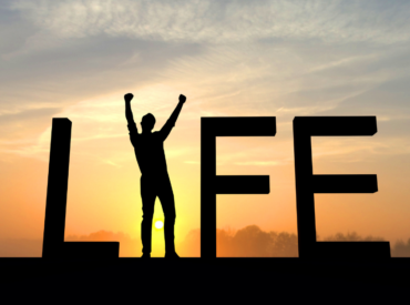 Silhouette of a person raising arms in front of large “LIFE” letters at sunrise, symbolizing motivation and success