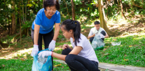 Volunteers wearing gloves collect plastic waste in a green park, placing bottles into a blue bag during a cleanup