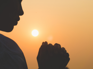 Silhouette of a person praying at sunset with hands clasped, symbolizing faith, spirituality, and peaceful reflection