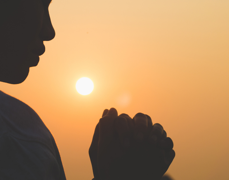 Silhouette of a person praying at sunset with hands clasped, symbolizing faith, spirituality, and peaceful reflection