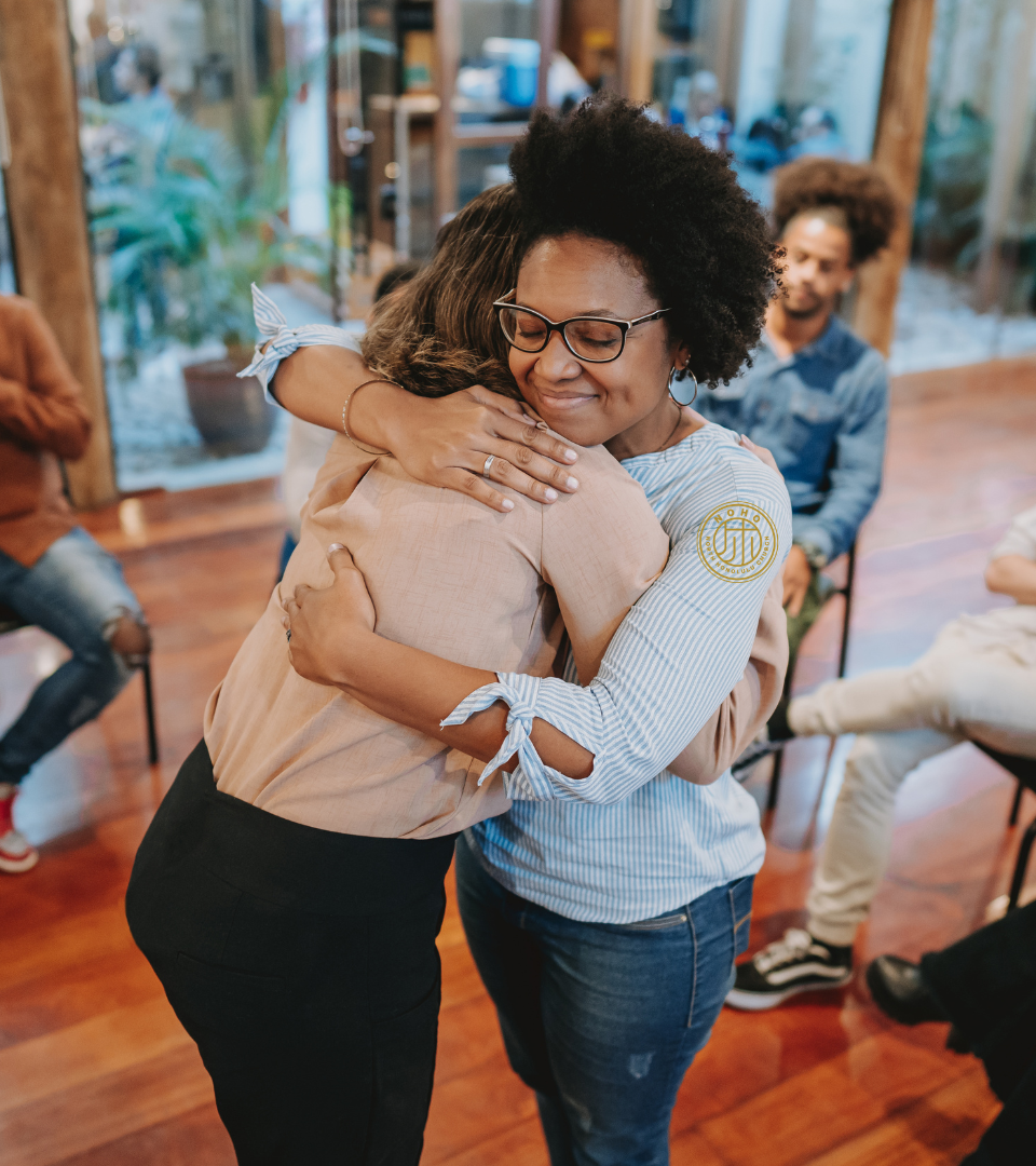 Two women sharing a warm hug in a group setting, showing emotional support, connection, and community bonding